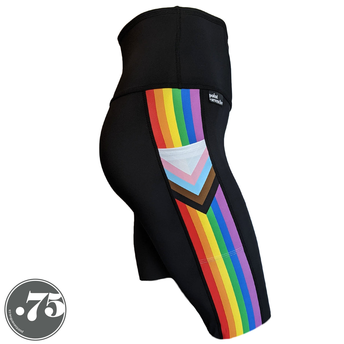 A pair of black spandex knee length shorts on a mannequin, the leggings have a 3.5” wide stripe down the side that has a printed fabric with the stripes of the Progress Pride Flag vertically and the are chevrons placed at the top of the pocket. The stripes are red, orange, yellow, green, blue and purple. The chevrons are white, pink, light blue, brown and black and point down from the top of the pocket.