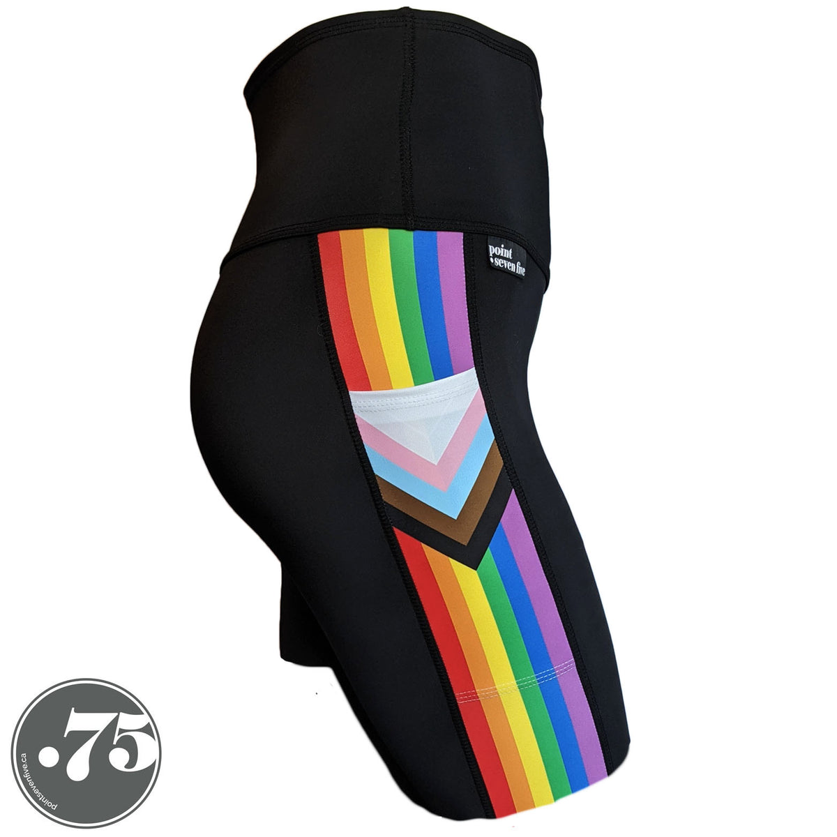 A pair of black spandex skate length shorts on a mannequin, the leggings have a 3.5” wide stripe down the side that has a printed fabric with the stripes of the Progress Pride Flag vertically and the are chevrons placed at the top of the pocket. The stripes are red, orange, yellow, green, blue and purple. The chevrons are white, pink, light blue, brown and black and point down from the top of the pocket.