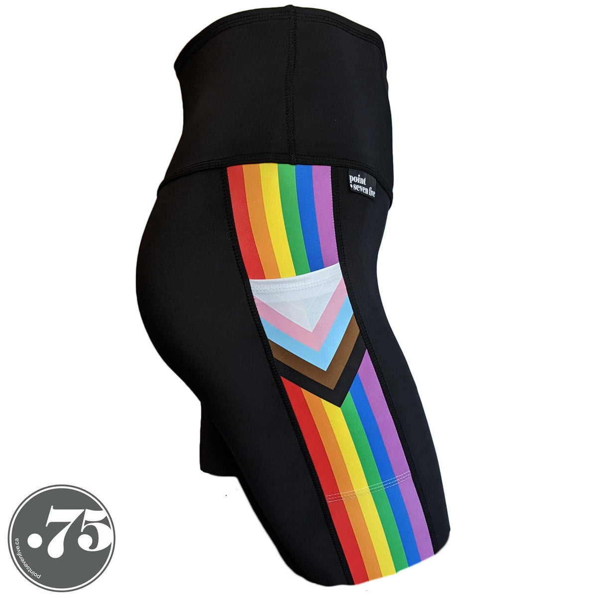 A pair of black spandex skate length shorts on a mannequin, the leggings have a 3.5” wide stripe down the side that has a printed fabric with the stripes of the Progress Pride Flag vertically and the are chevrons placed at the top of the pocket. The stripes are red, orange, yellow, green, blue and purple. The chevrons are white, pink, light blue, brown and black and point down from the top of the pocket.