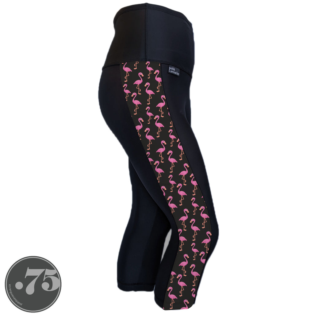 A pair of black spandex capri leggings against a white background. The leggings are calf length and have a panel on the side with a pocket, the panel has fabric with a charcoal grey background with scattered cartoon pink flamingos on it.