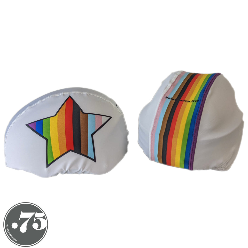 Two white helmet covers on two different helmets. The left cover has a star on the side with stripes in the colours of the inclusive pride flag. The right cover has a stripe down the middle with stripes in the colours of the inclusive pride flag. 